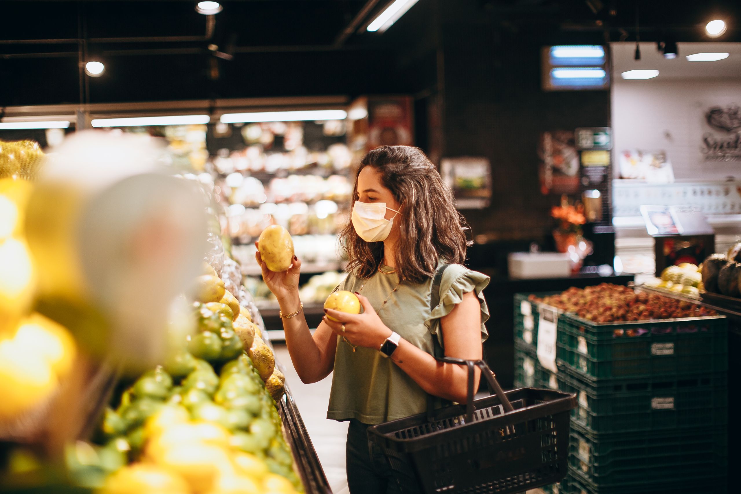 A woman wearing a face mask shops for fresh produce in a well-lit grocery store, holding fruits.
