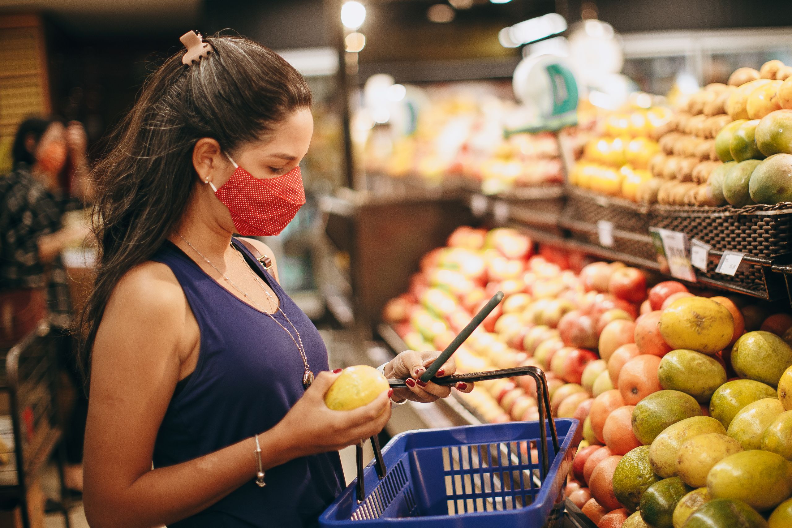A woman wearing a mask selects fresh fruits in a supermarket produce aisle.