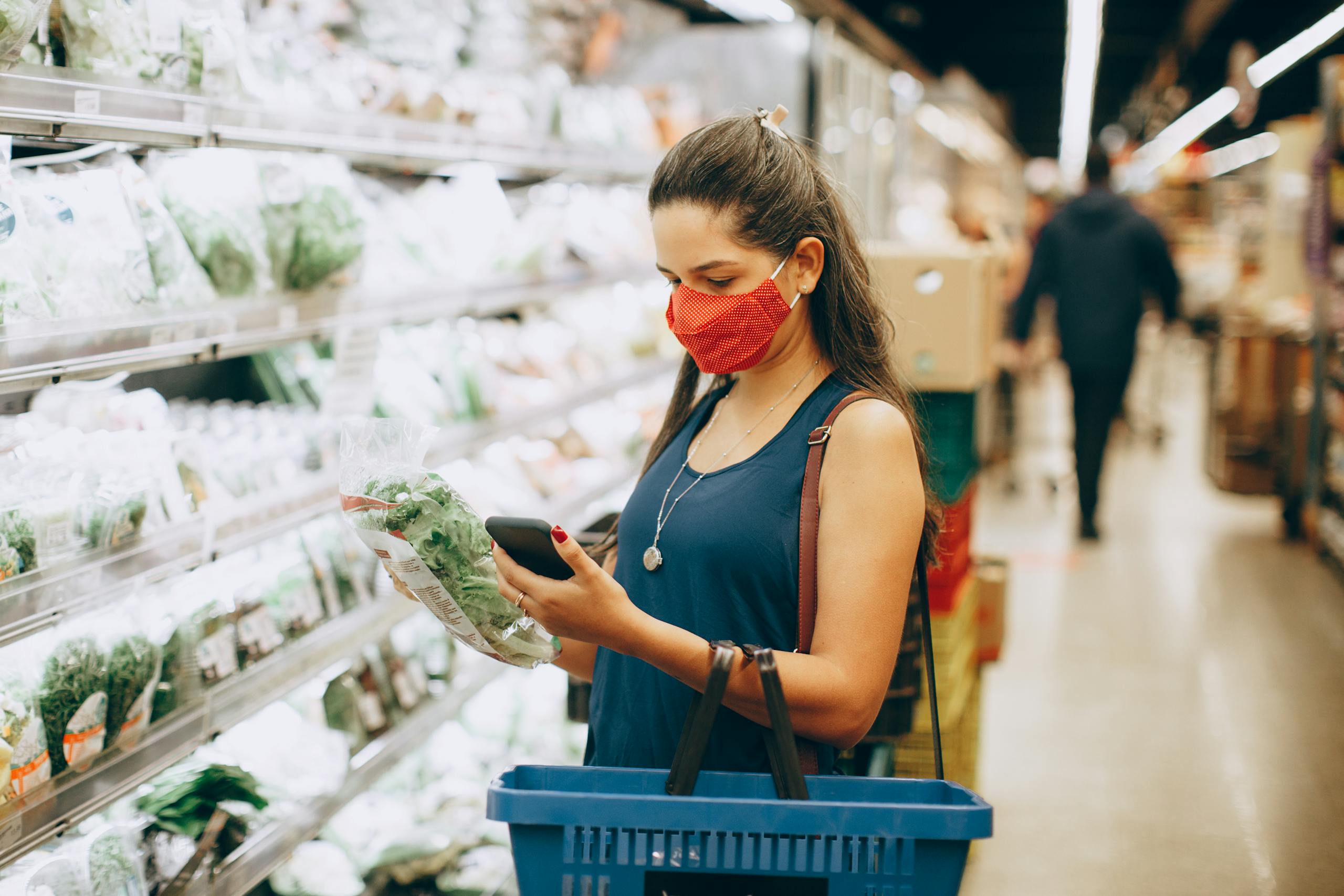 A woman wearing a mask shops for groceries in a supermarket, checking products with her phone.
