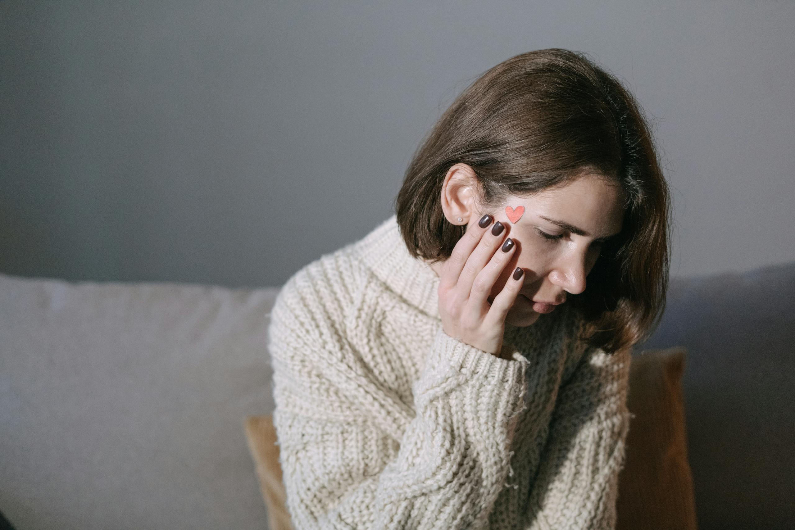 A woman wearing a sweater rests her hand on her face, displaying a heart sticker, in a softly lit room.