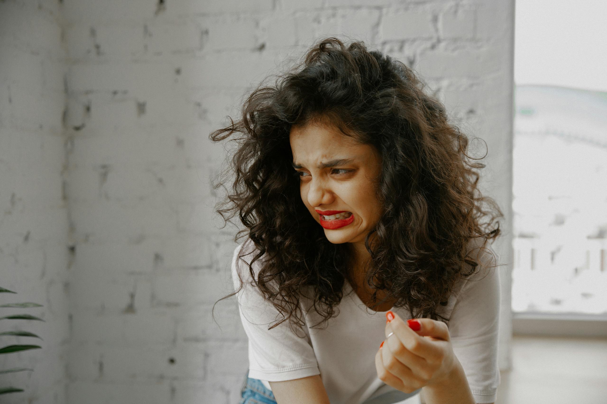A woman with curly hair and red lips expresses a disgusted facial expression indoors.