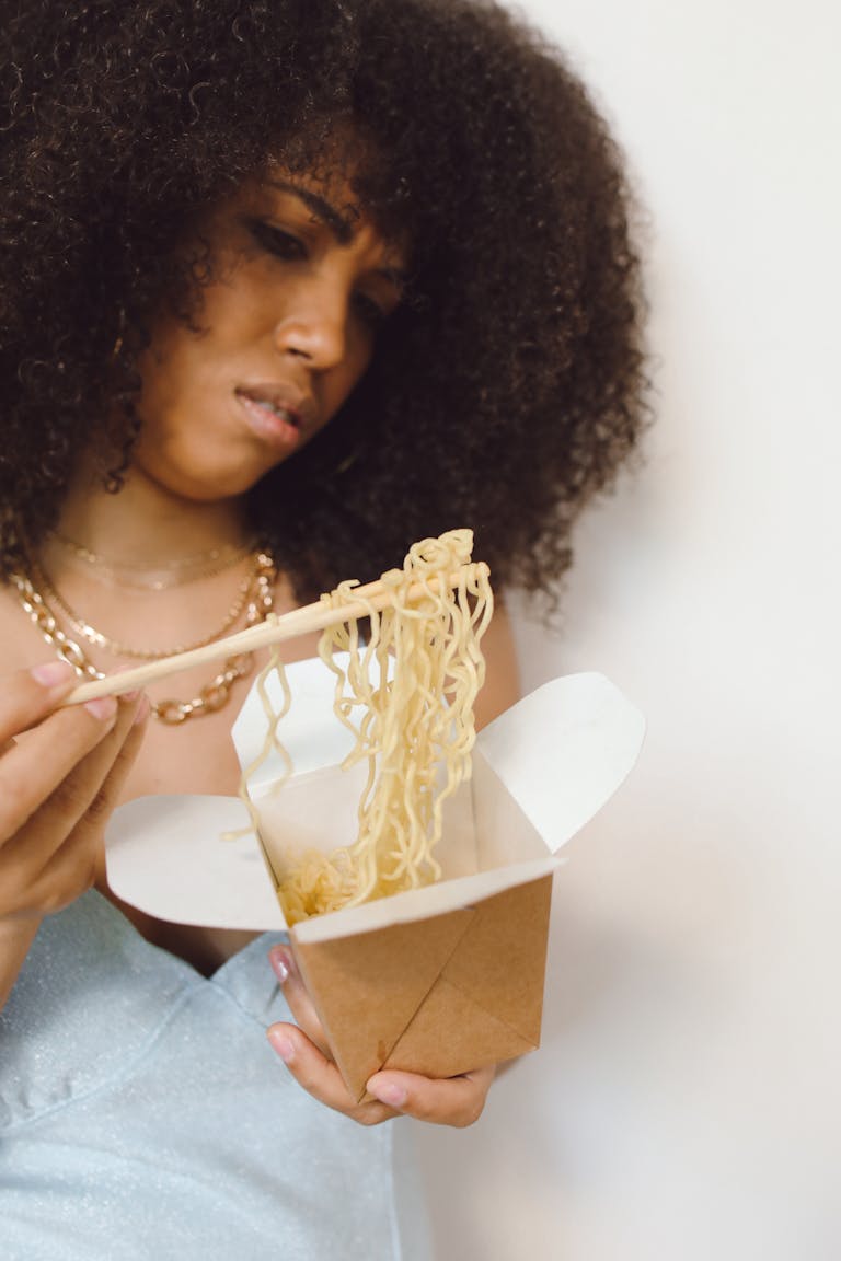 A woman with curly hair examines noodles in a takeout box, showing a thoughtful expression.