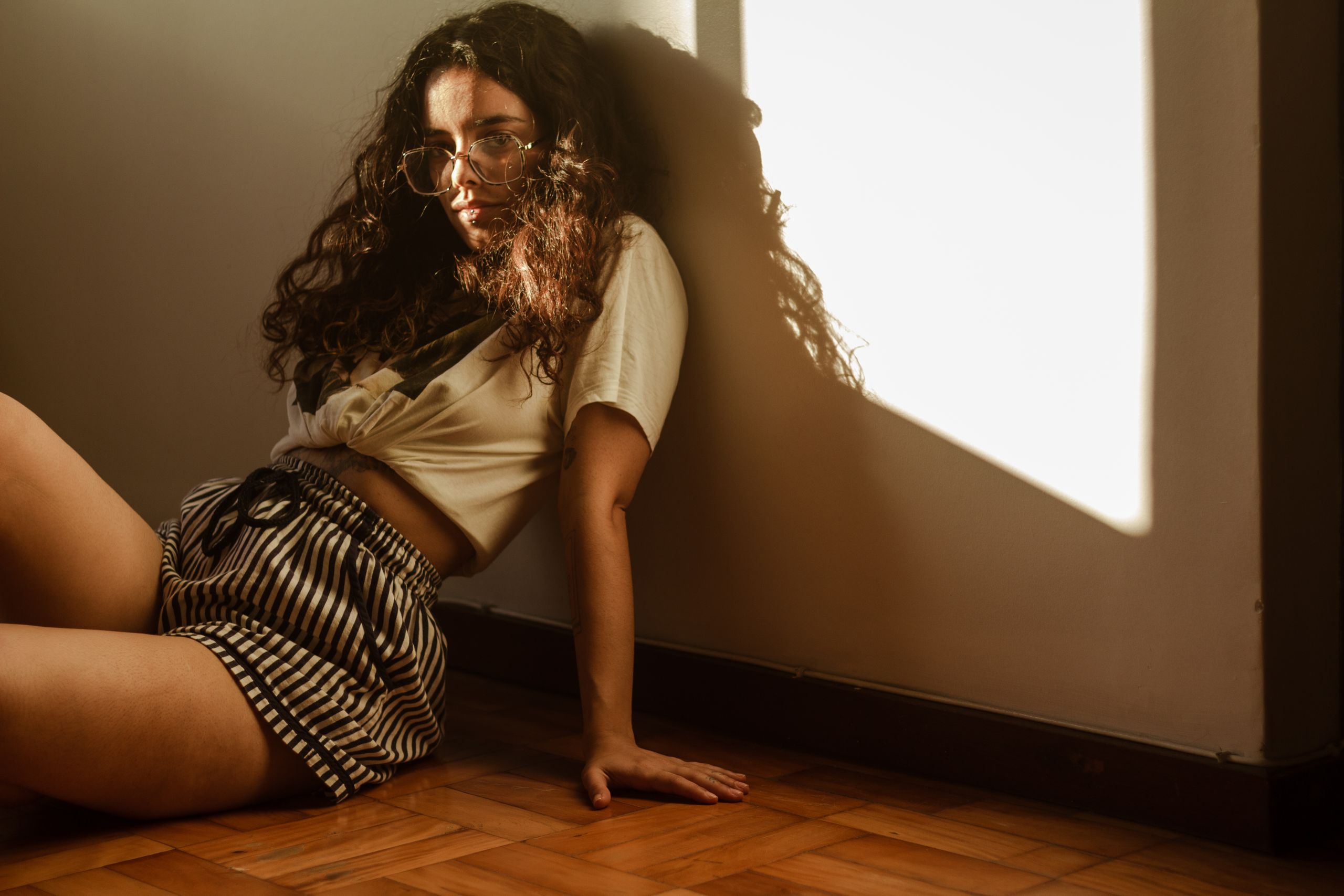 A woman with curly hair sits on a wooden floor, illuminated by warm sunlight, creating a relaxed atmosphere.
