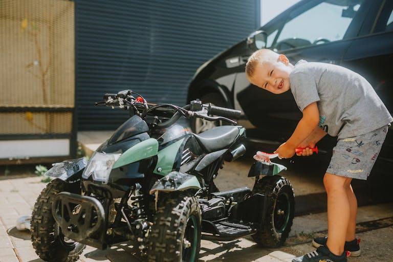 A young boy cheerfully cleans a kids' ATV outdoors on a sunny day, showcasing a playful moment.