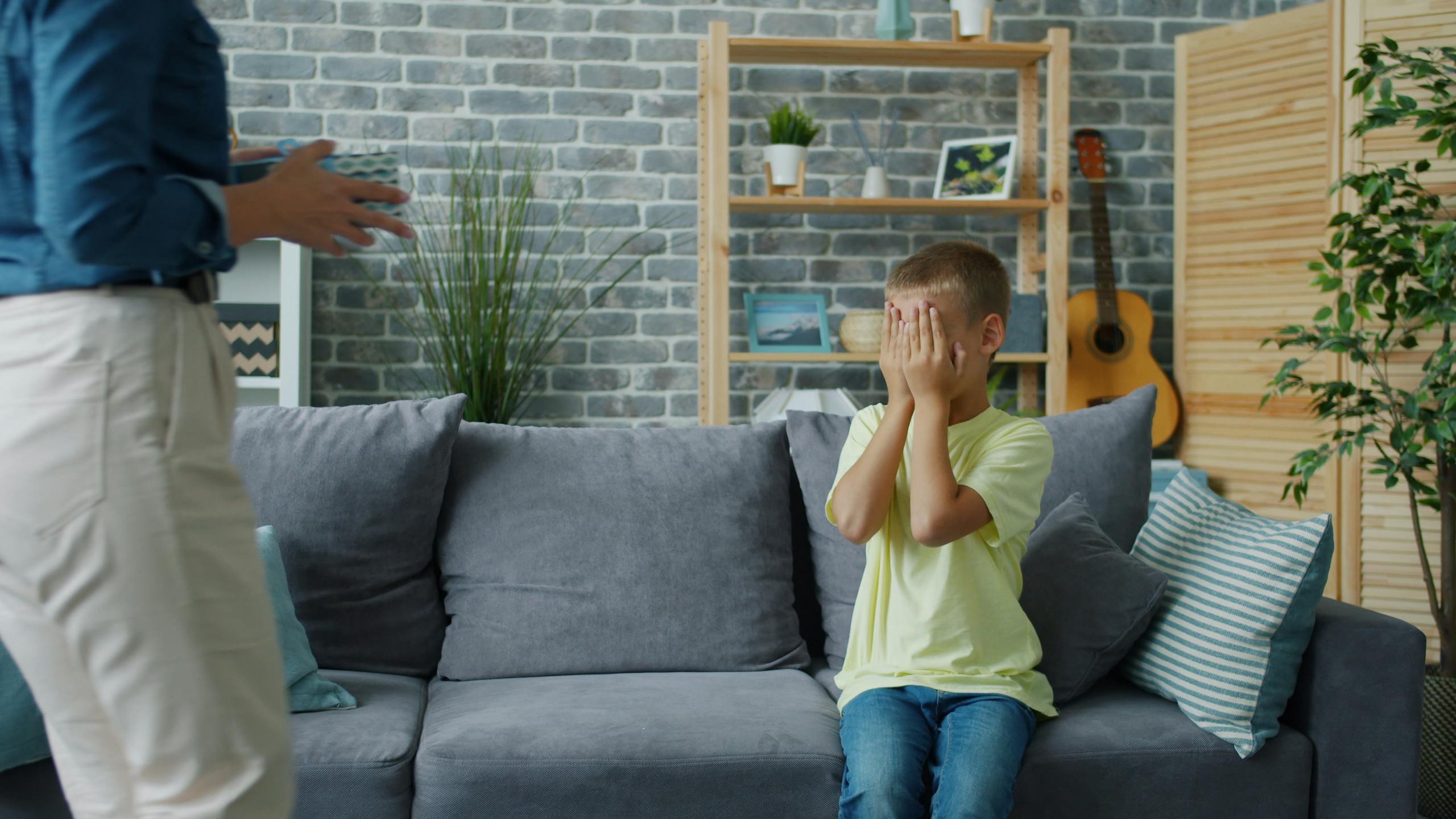 A young boy covers his eyes on a sofa in a cozy living room setting.