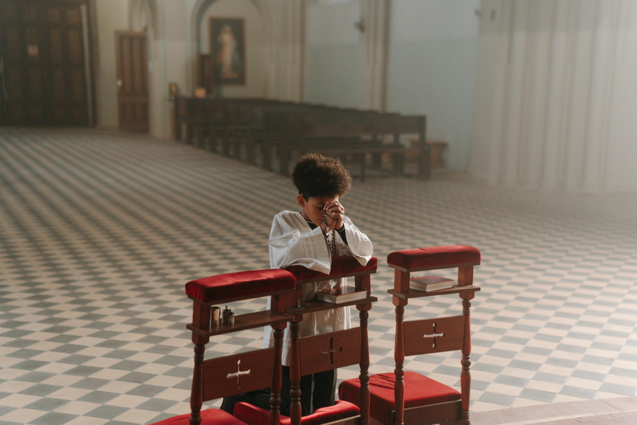 A young boy with curly hair is kneeling in prayer at an empty church, emphasizing solitude and faith.