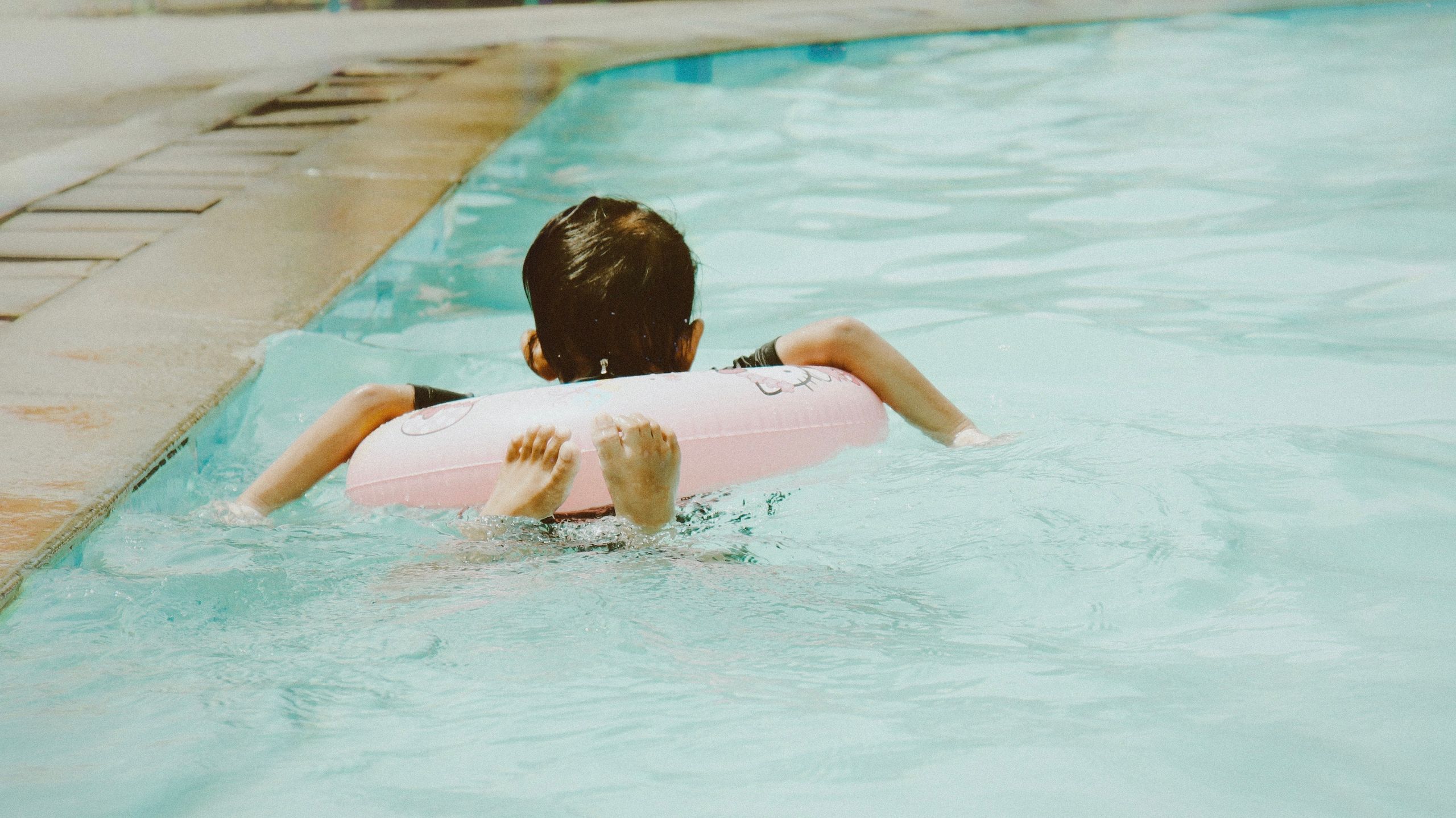 A young child swimming in a pool using a pink float, enjoying a sunny day.
