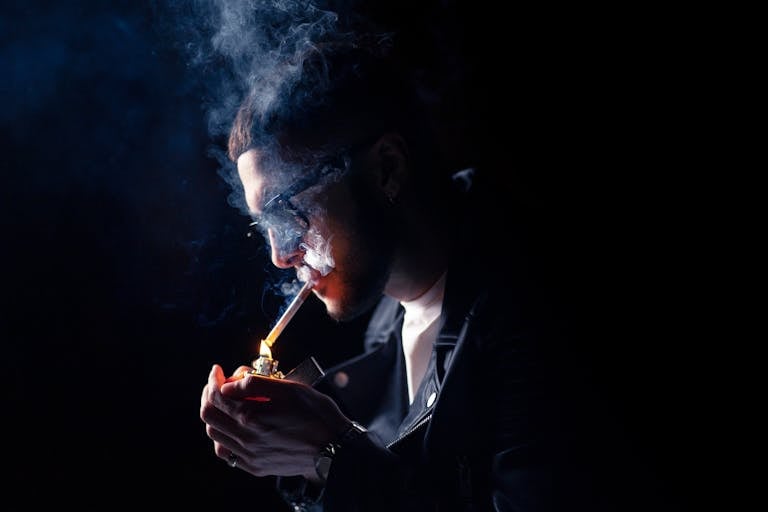 A young man in a leather jacket lighting a cigarette, surrounded by smoke.