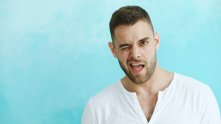 A young man playfully winks with a cheerful expression against a blue background.