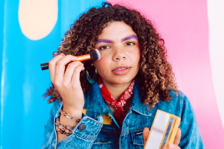 A young woman with curly hair applies makeup using a brush against a vibrant, colorful background.