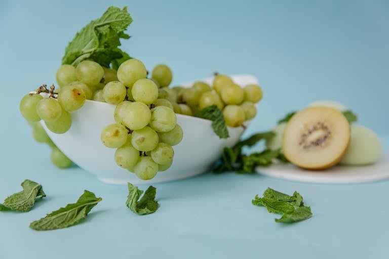 Aesthetic arrangement of green grapes with kiwi and mint leaves on a light blue background.