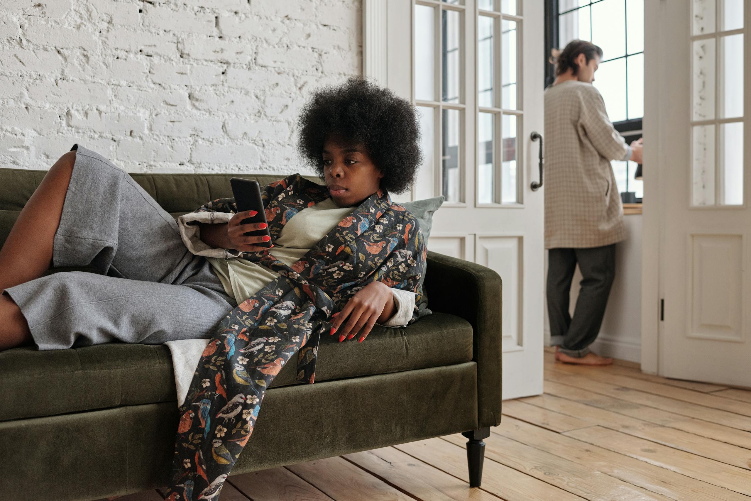 African American woman lounging on a sofa, texting, while man stands by window.