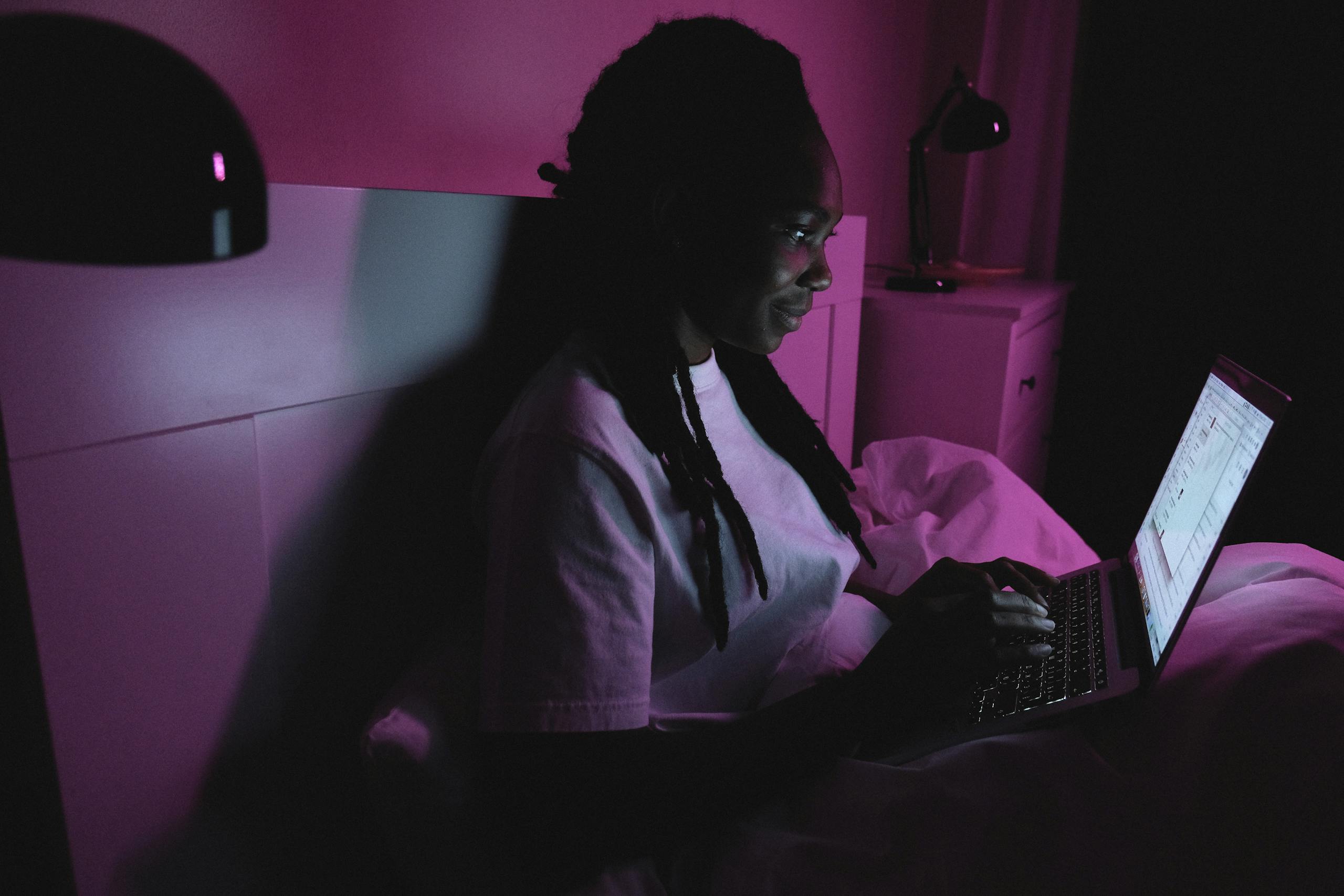 An adult woman using a laptop in bed under purple lighting, representing modern remote work at home.