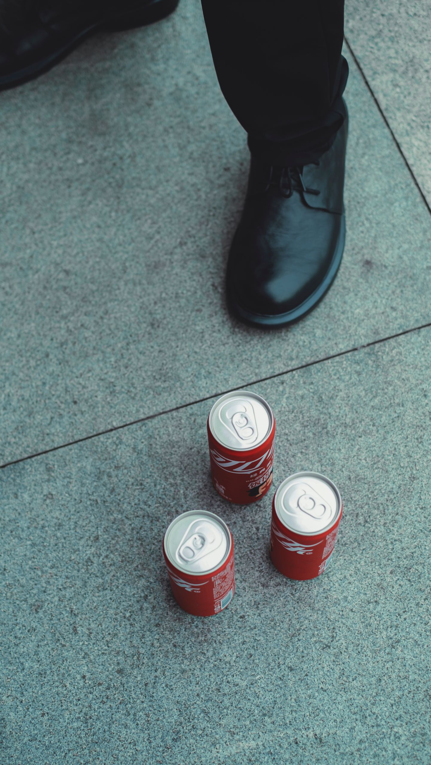 An artistic shot of three soda cans placed on a pavement near a black shoe, creating a modern urban vibe.