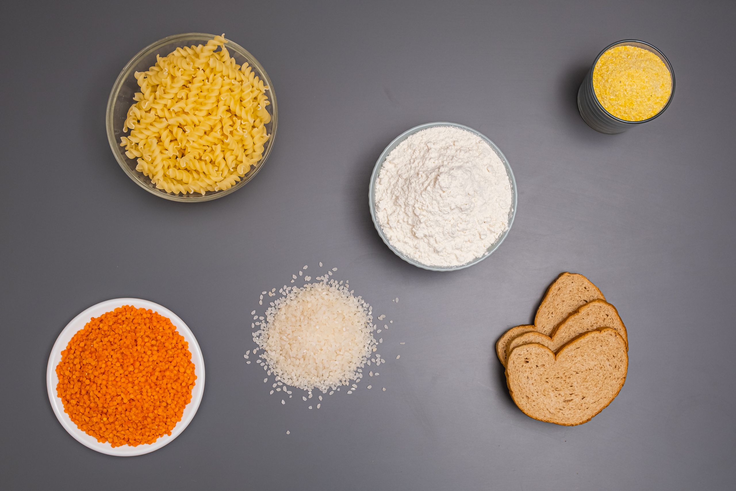 An assortment of dry ingredients including pasta, rice, and bread displayed on a grey background.