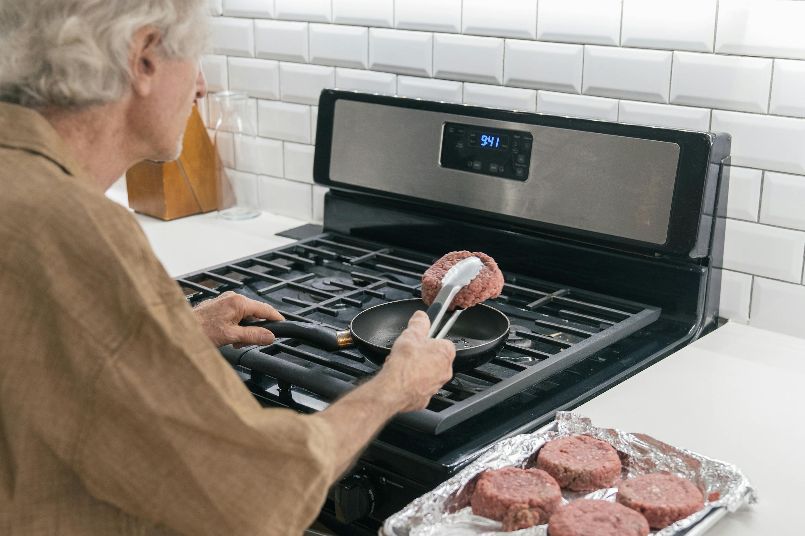 An elderly man prepares beef patties in a kitchen using a non-stick pan.