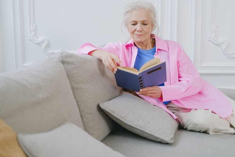 An elderly woman in a pink shirt enjoys relaxing on a couch while reading a book indoors.