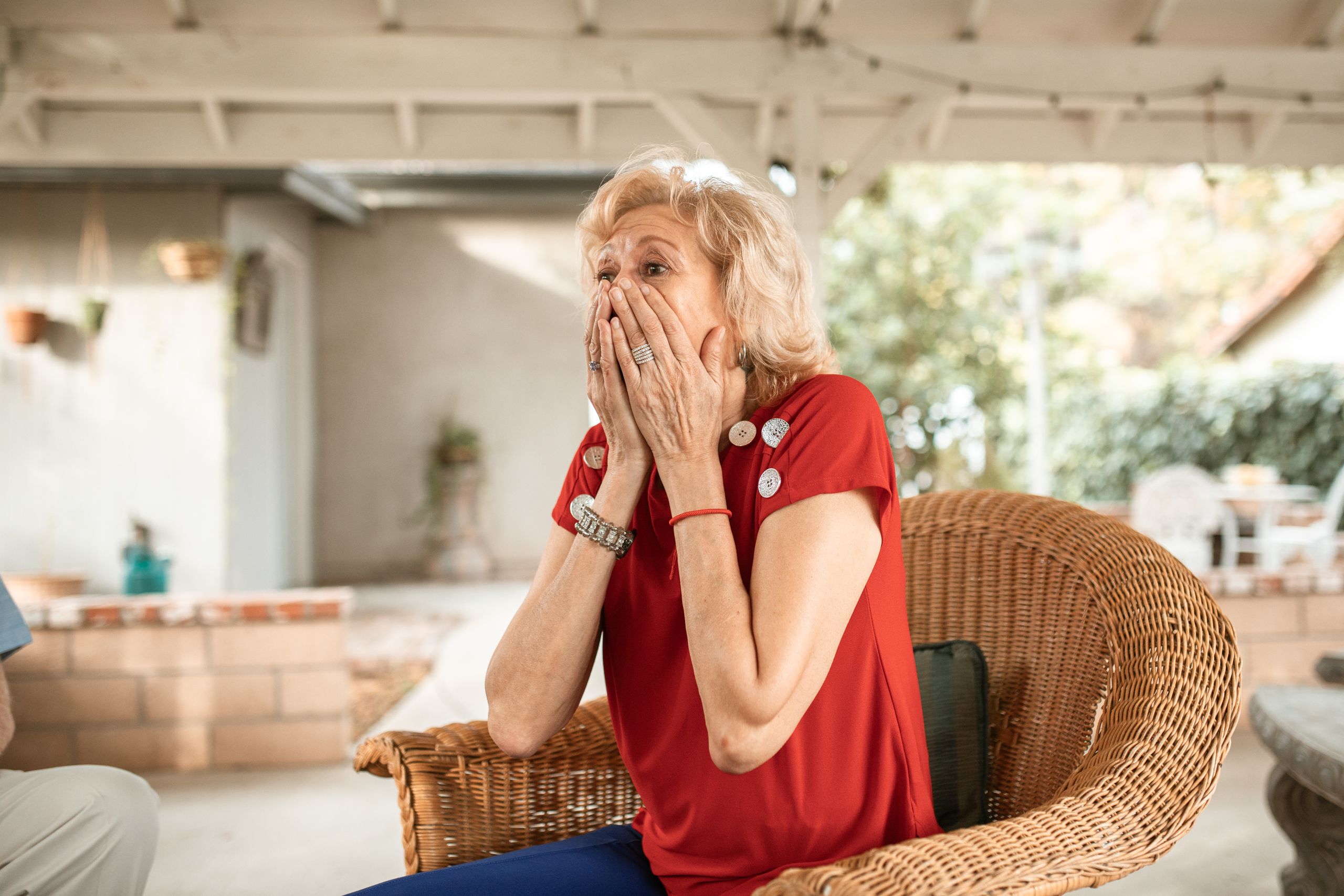 An elderly woman with a surprised expression sits in a wicker armchair indoors.