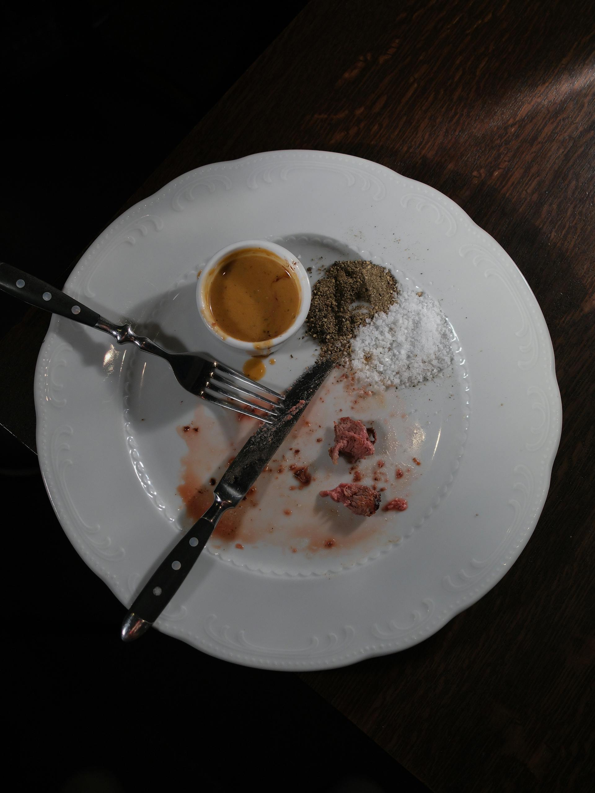 An empty dinner plate with steak remnants, cutlery, and seasonings on a dark table.