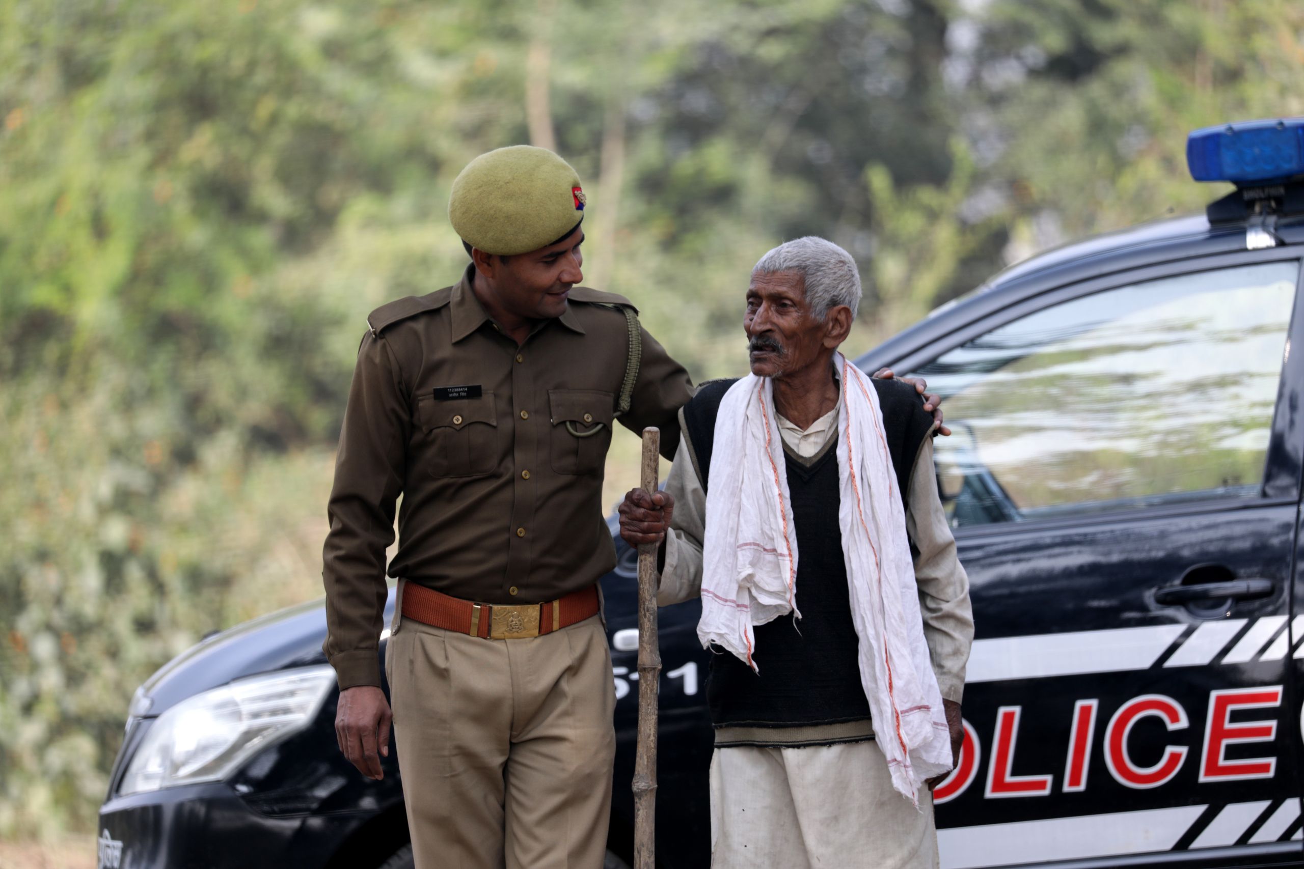 An Indian policeman assists an elderly man with a walking stick next to a police car outdoors.