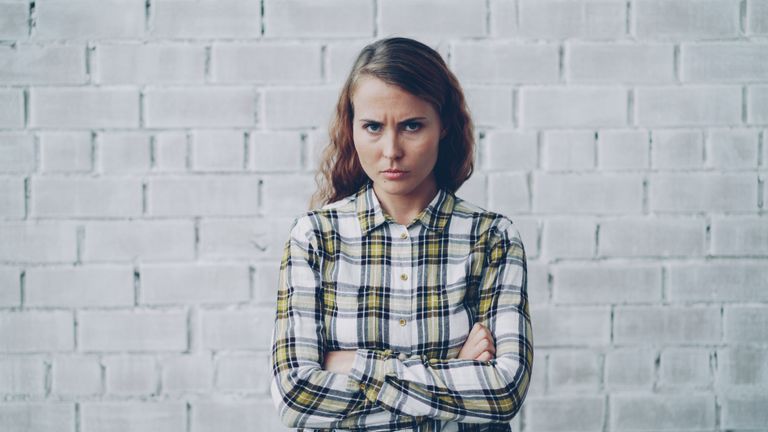 Angry woman with arms crossed in plaid shirt standing against a brick wall.