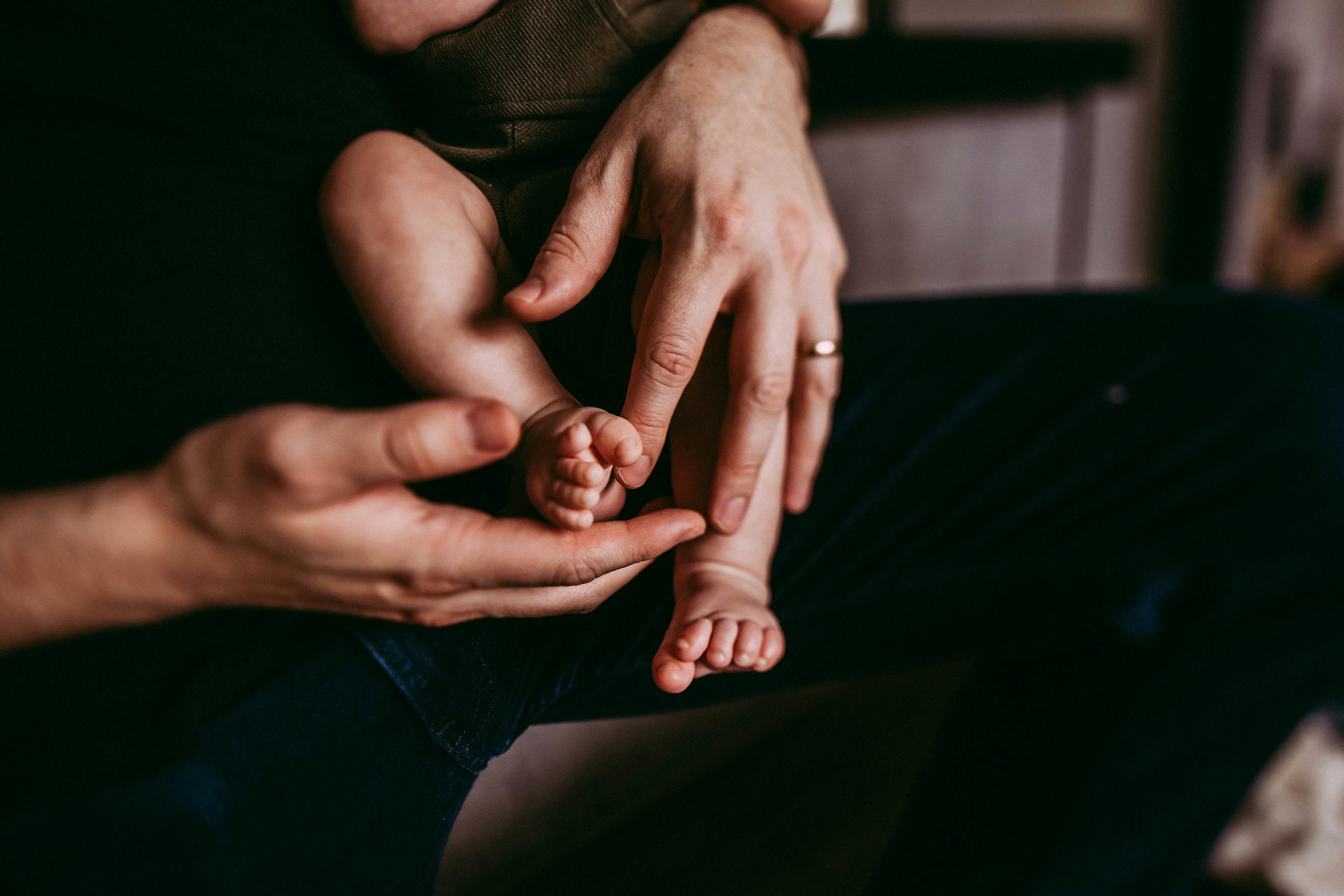 Anonymous caring parent with adorable barefoot little baby in arms standing in light living room on blurred background at home