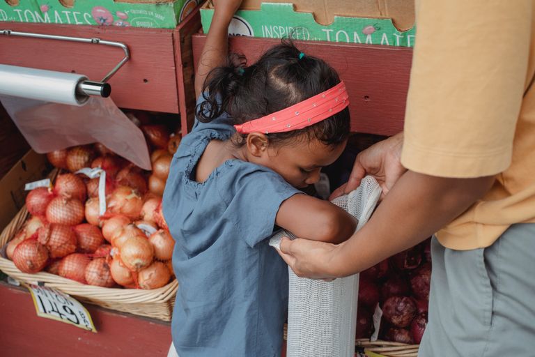 Asian girl and mother putting vegetables in bag while doing groceries in local street market and standing near stall