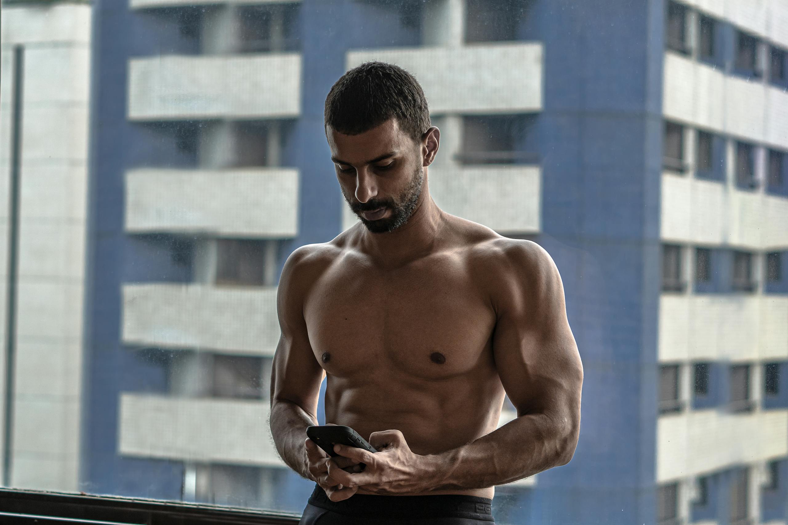 Athletic shirtless man using smartphone by a window in an urban setting.