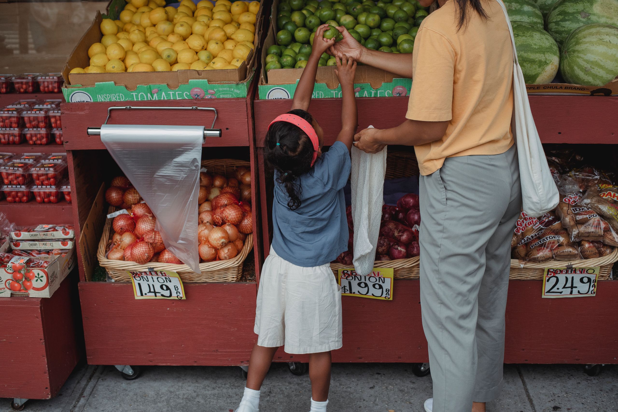 Back view of anonymous ethnic small girl choosing limes from box standing near mother with reusable bag
