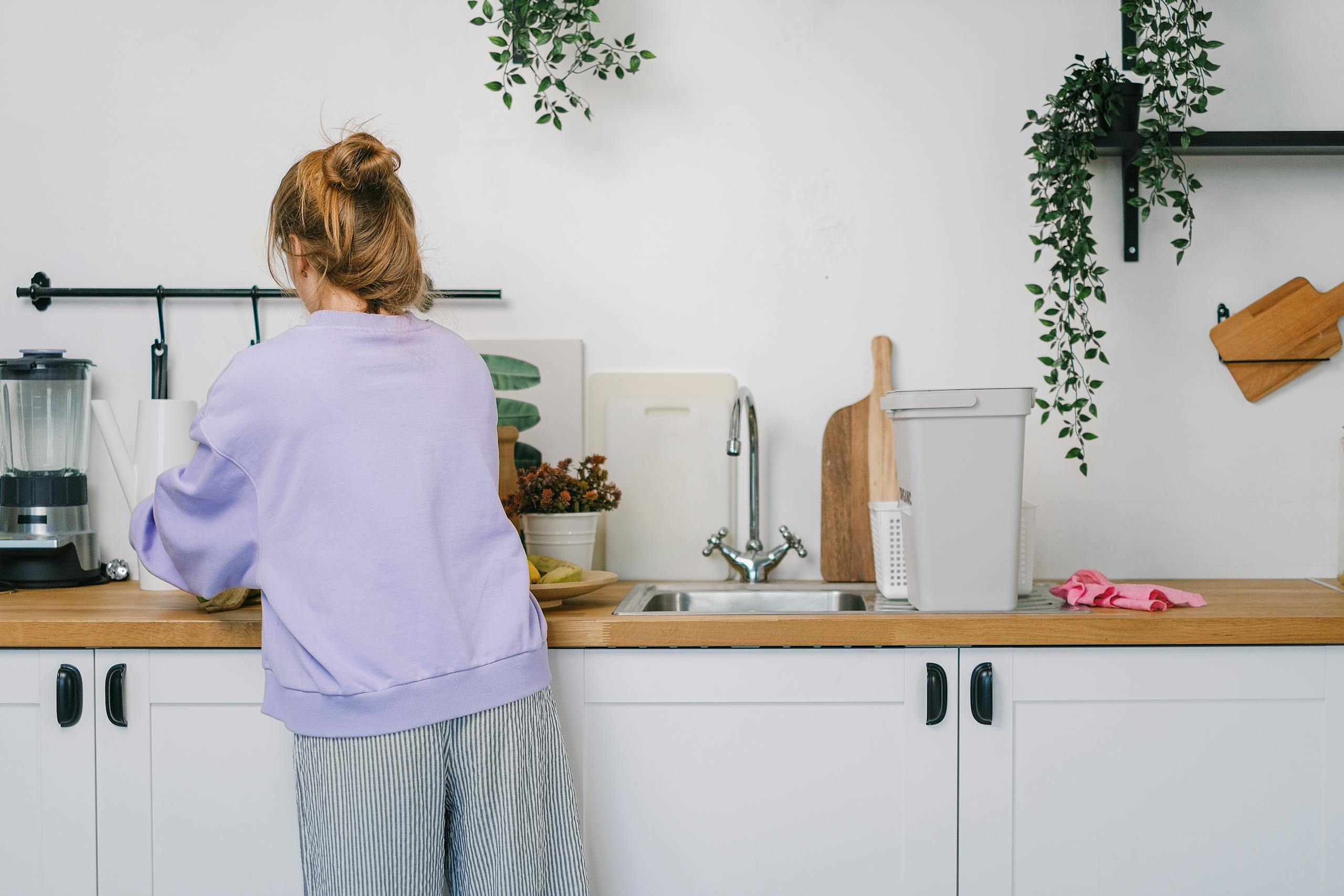 Back view of unrecognizable young female in casual clothes standing at wooden counter in light kitchen in daytime
