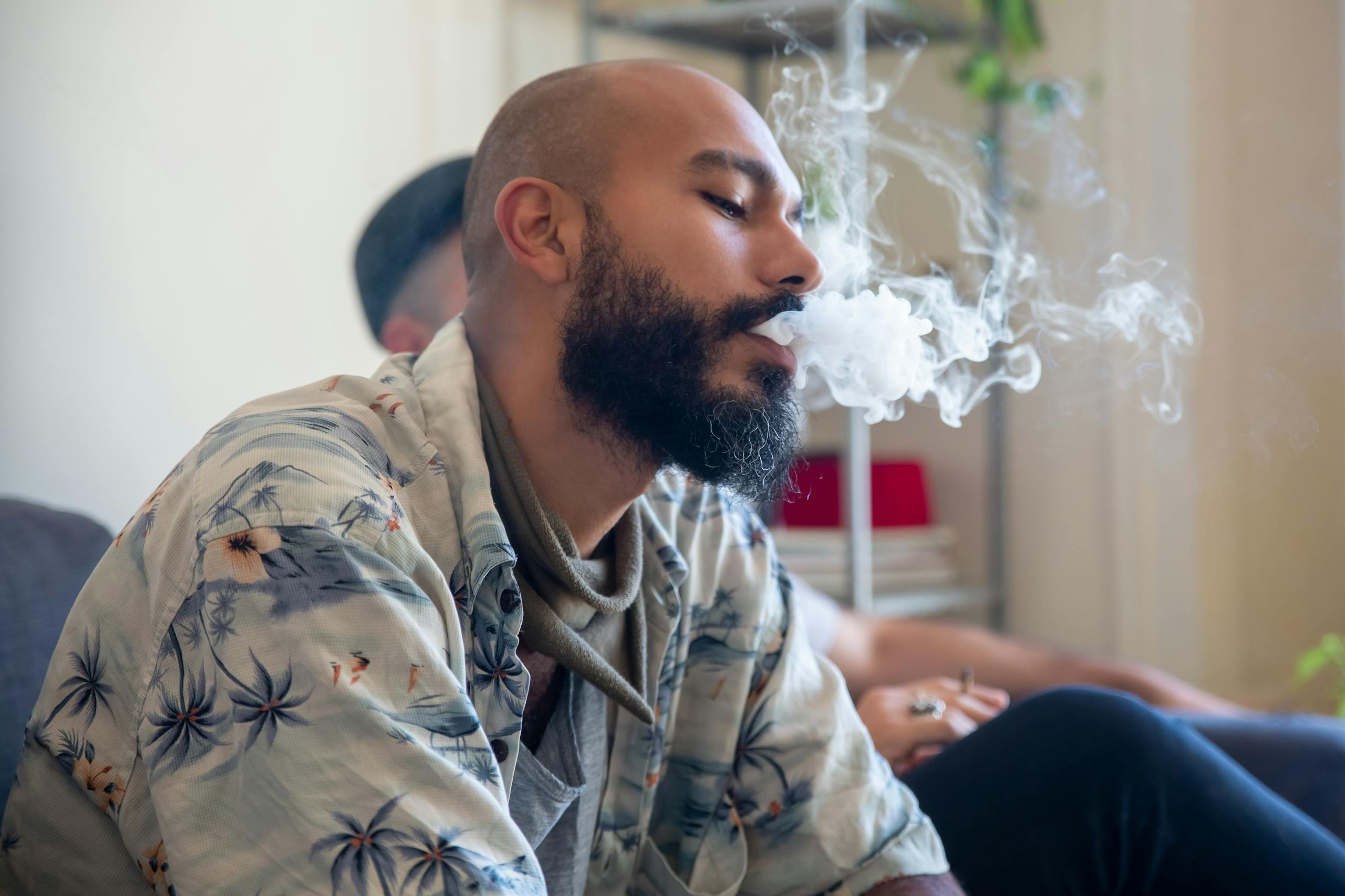 Bald bearded man exhaling smoke indoors, capturing a relaxed moment with a tropical shirt.
