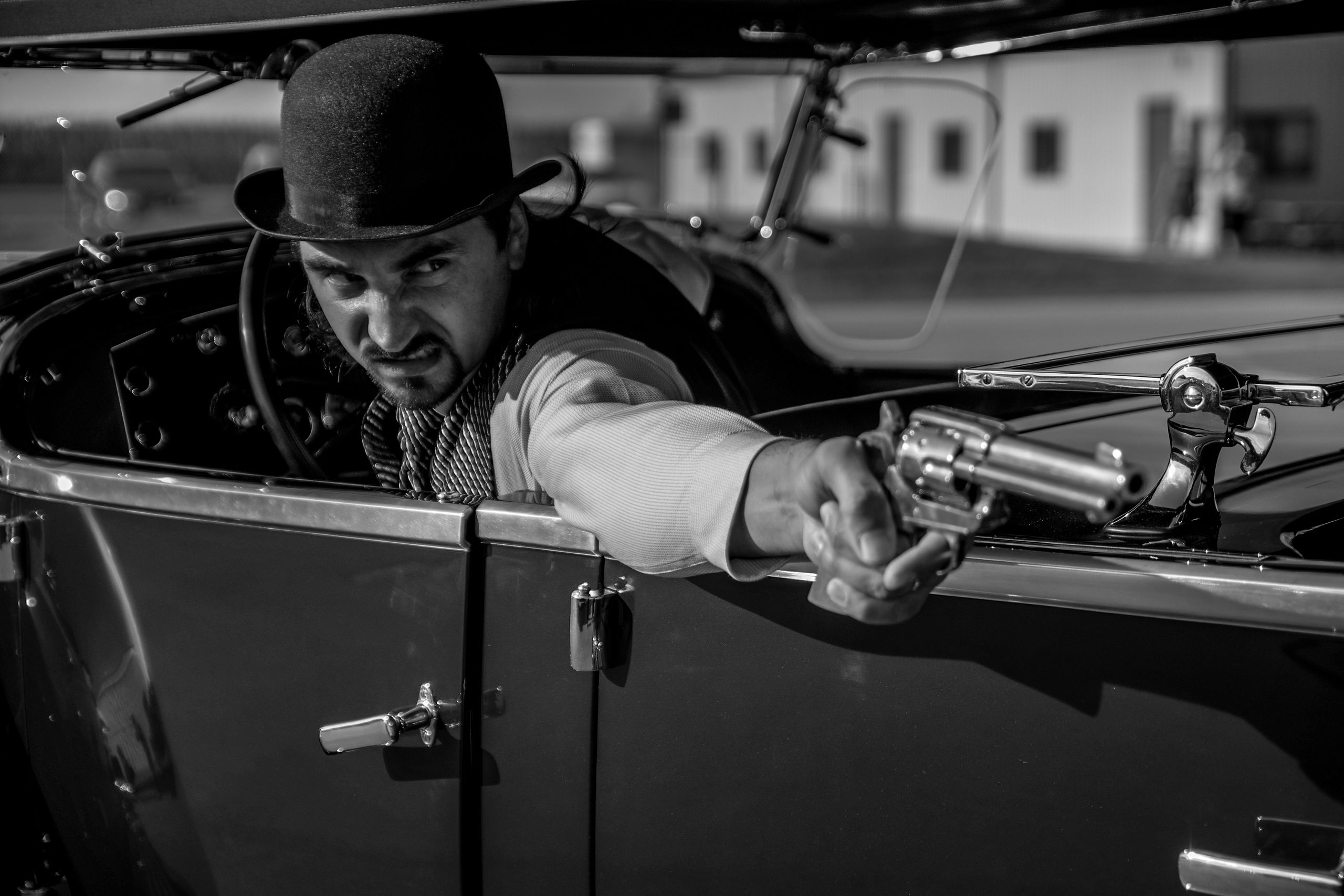 Black and white photo of a man in vintage attire aiming a revolver from a classic car.