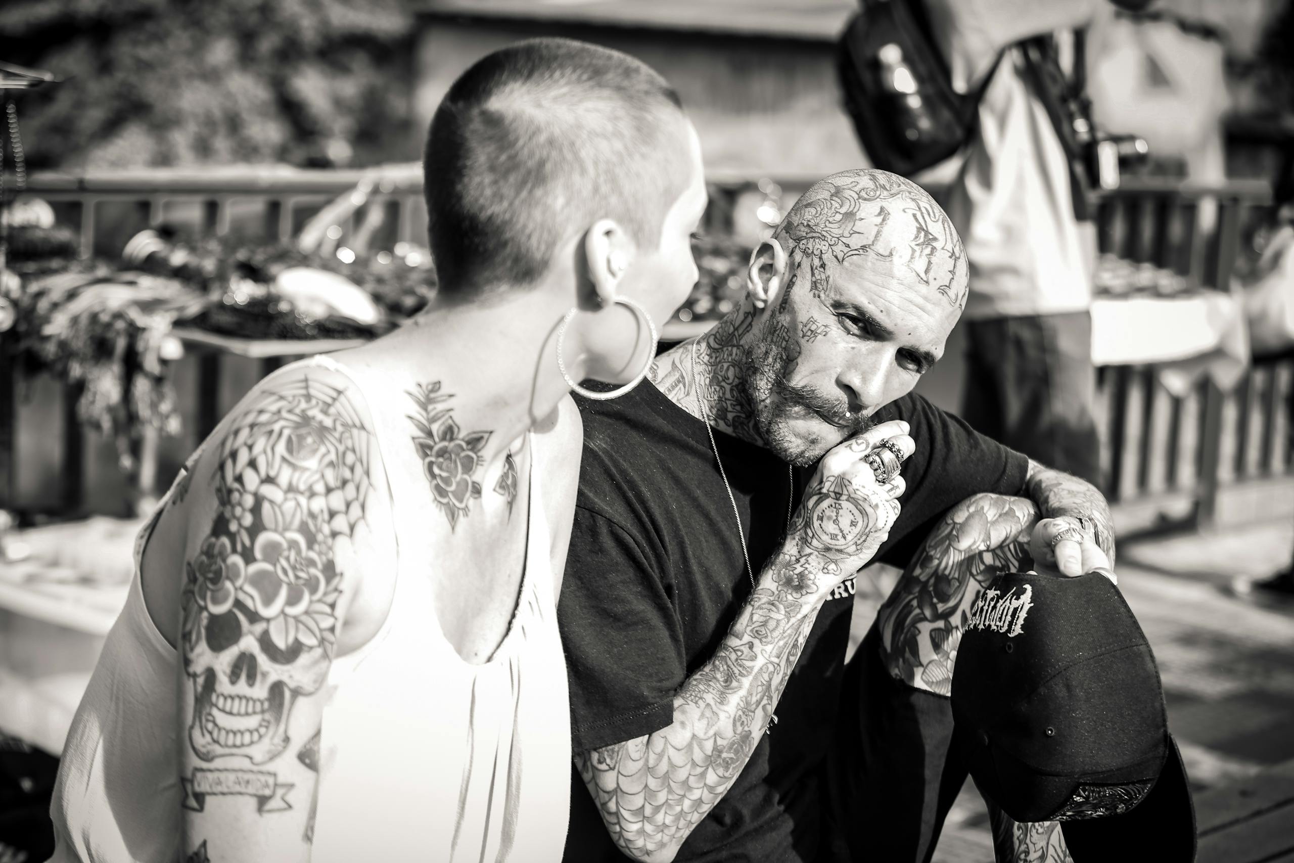 Black and white photo of tattooed adults conversing outdoors. Focus on tattoos and personal style.