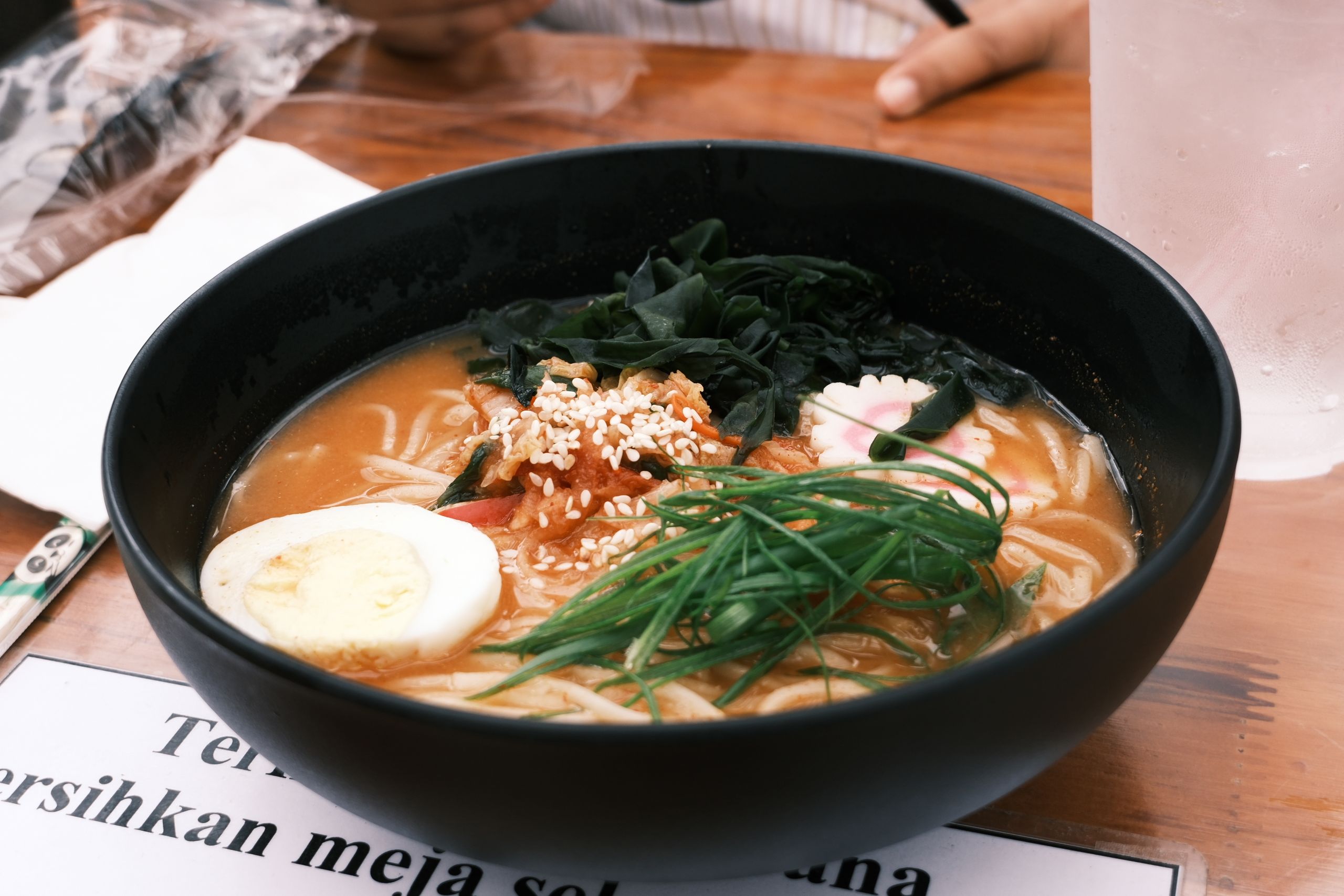 Bowl of ramen with egg, seaweed, green onions, and sesame seeds in an Asian restaurant setting.