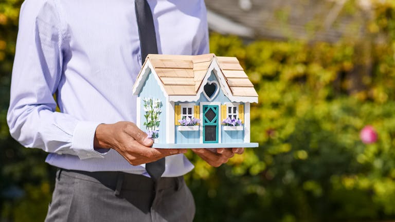 A businessman holds a small house model outdoors, symbolizing real estate investment.