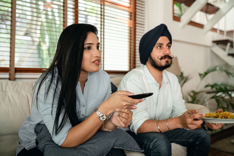 Calm Indian woman with remote control and man in turban and with mustache having meal while watching TV on couch