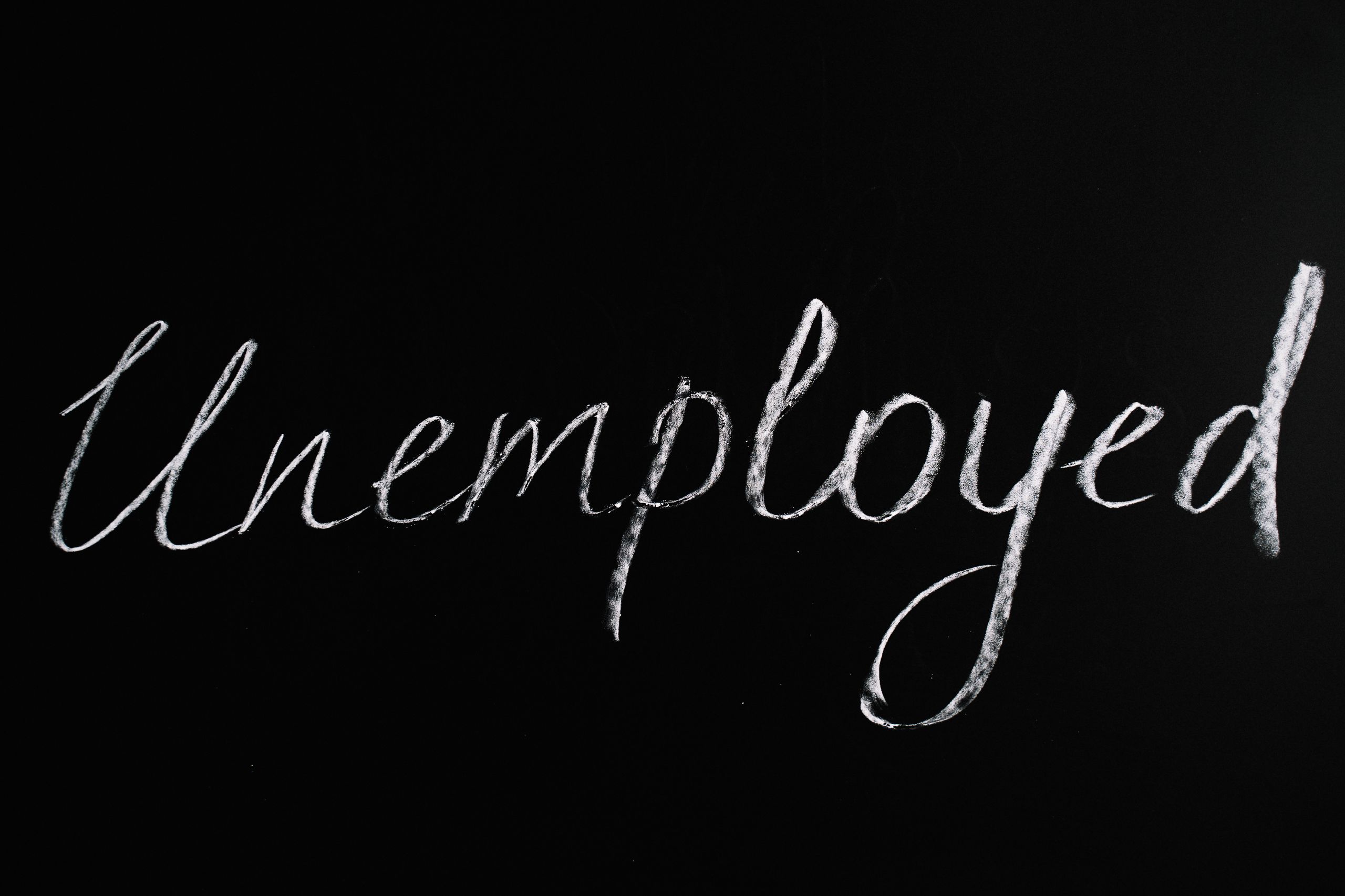 Chalk cursive text 'Unemployed' on black background, pointing to joblessness concept.