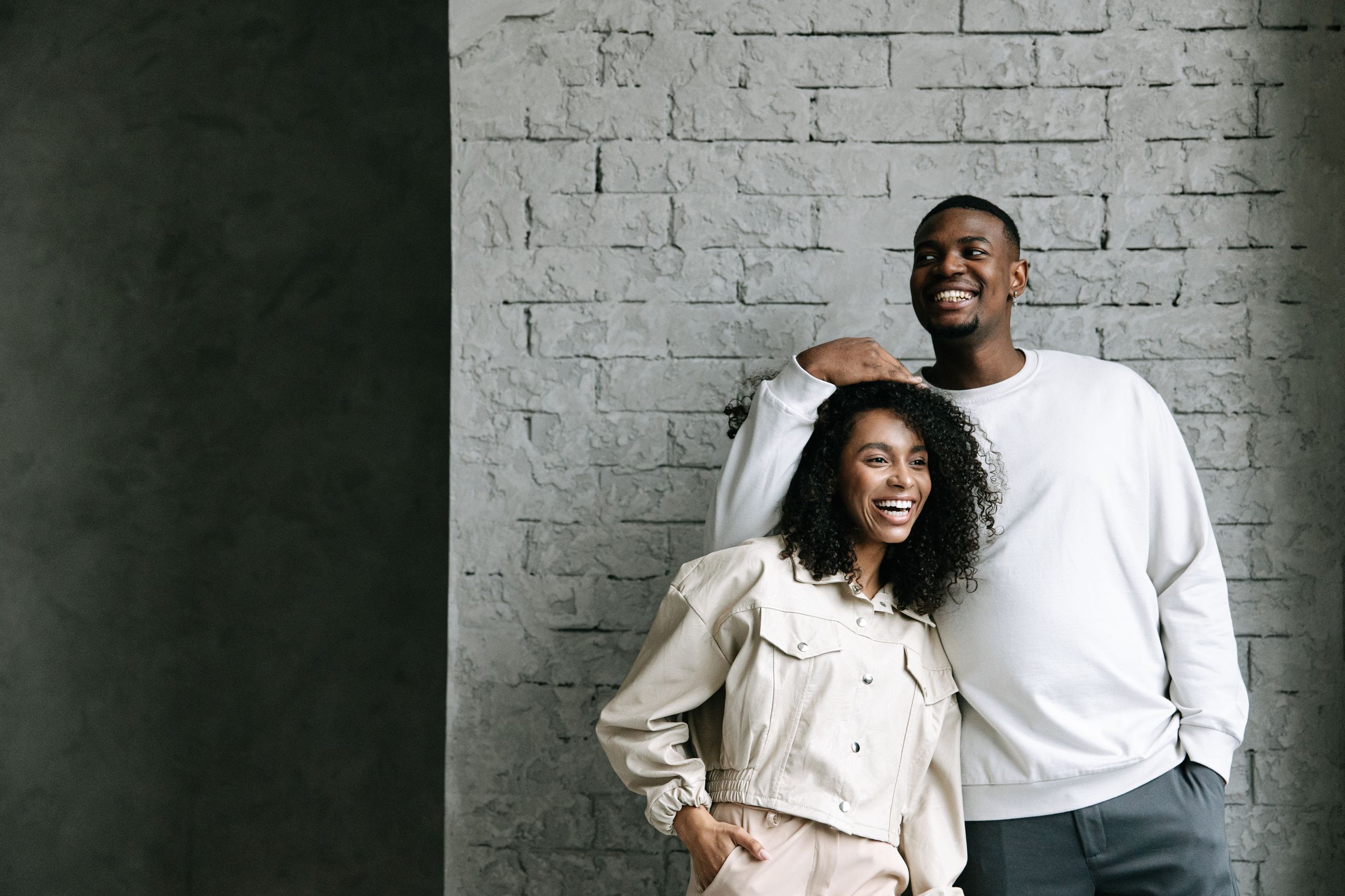 Cheerful couple standing and laughing against a modern brick wall, expressing happiness and style.