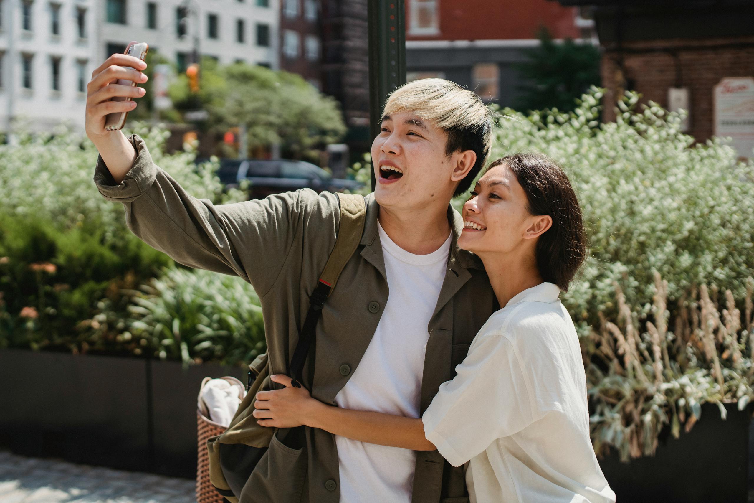 Cheerful young Asian couple in stylish clothes cuddling and smiling while taking selfie on mobile phone during date in city