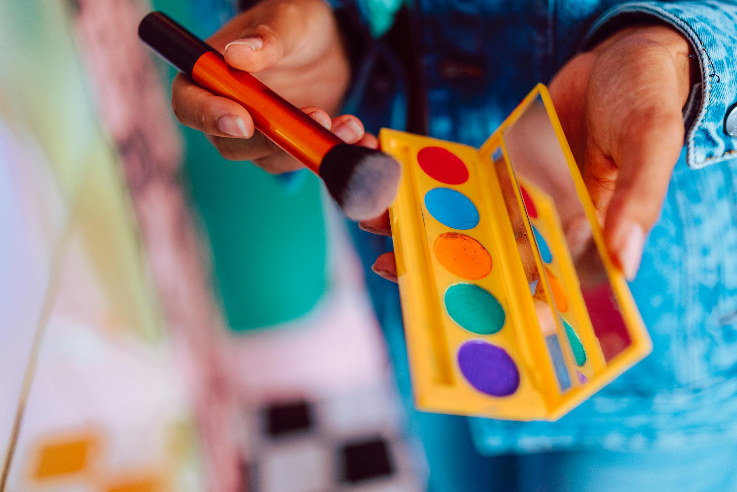 Close-up of a colorful eyeshadow palette and makeup brush held by a person.