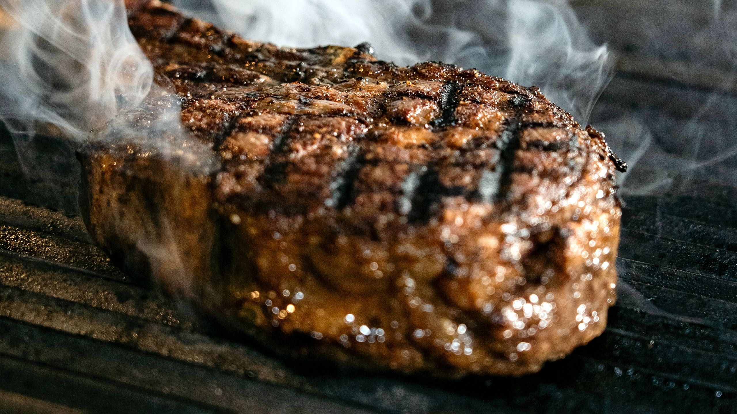 Close-up of a grilled steak with smoke rising, highlighting its juicy texture and grill marks.