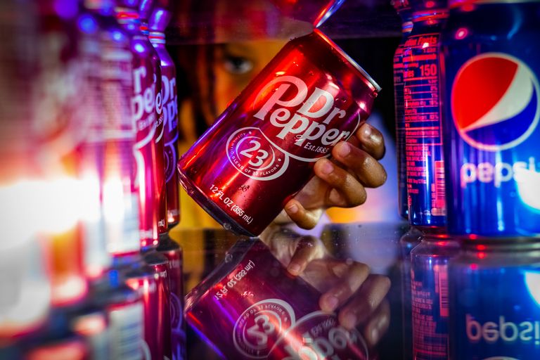 Close-up of a hand taking a Dr Pepper can from a refrigerator with colorful reflections.