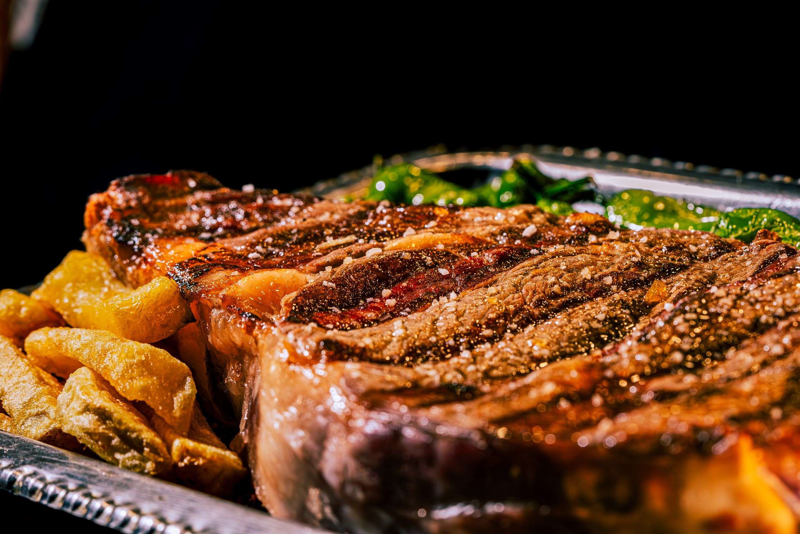 Close-up of a juicy grilled ribeye steak with pepper and sides on a silver platter.