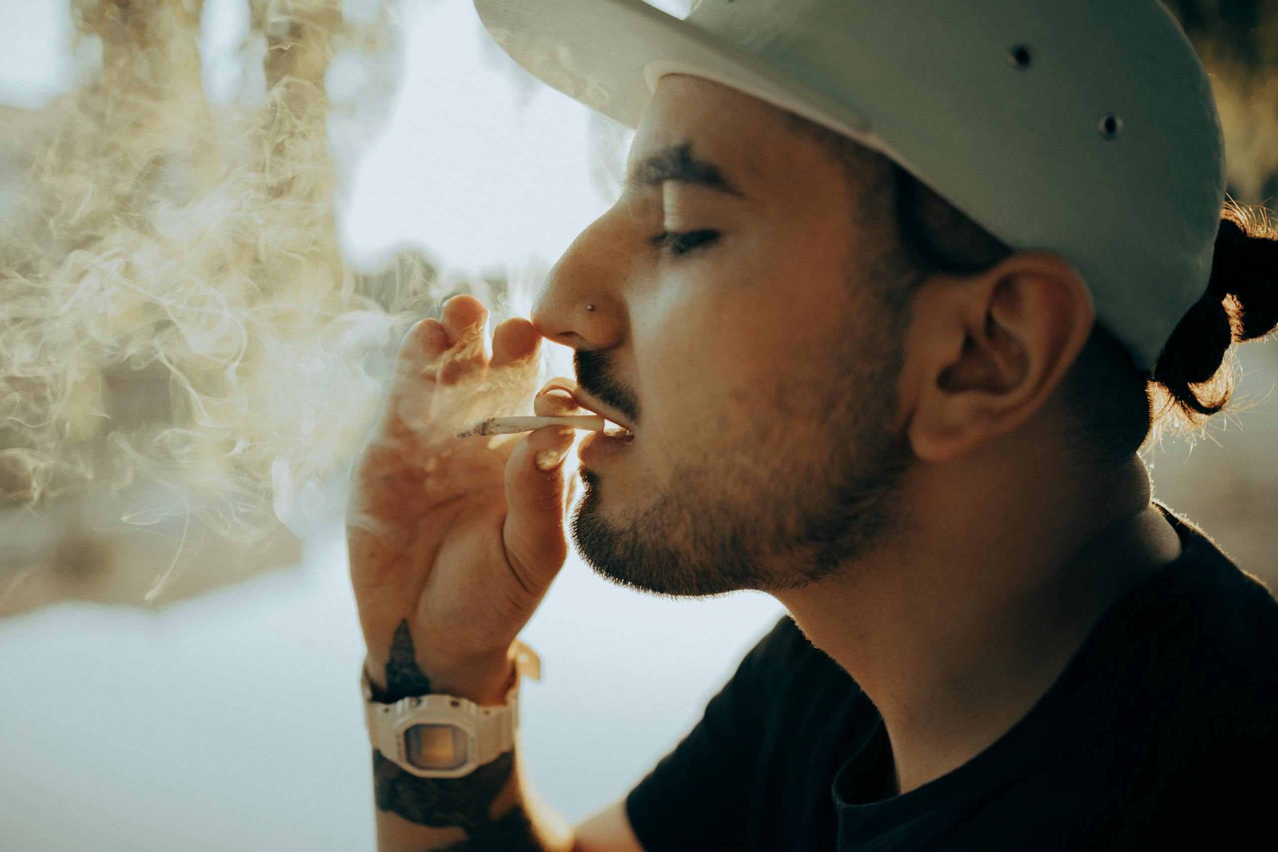 Close-up of a man with facial hair and cap smoking outdoors in a casual setting.