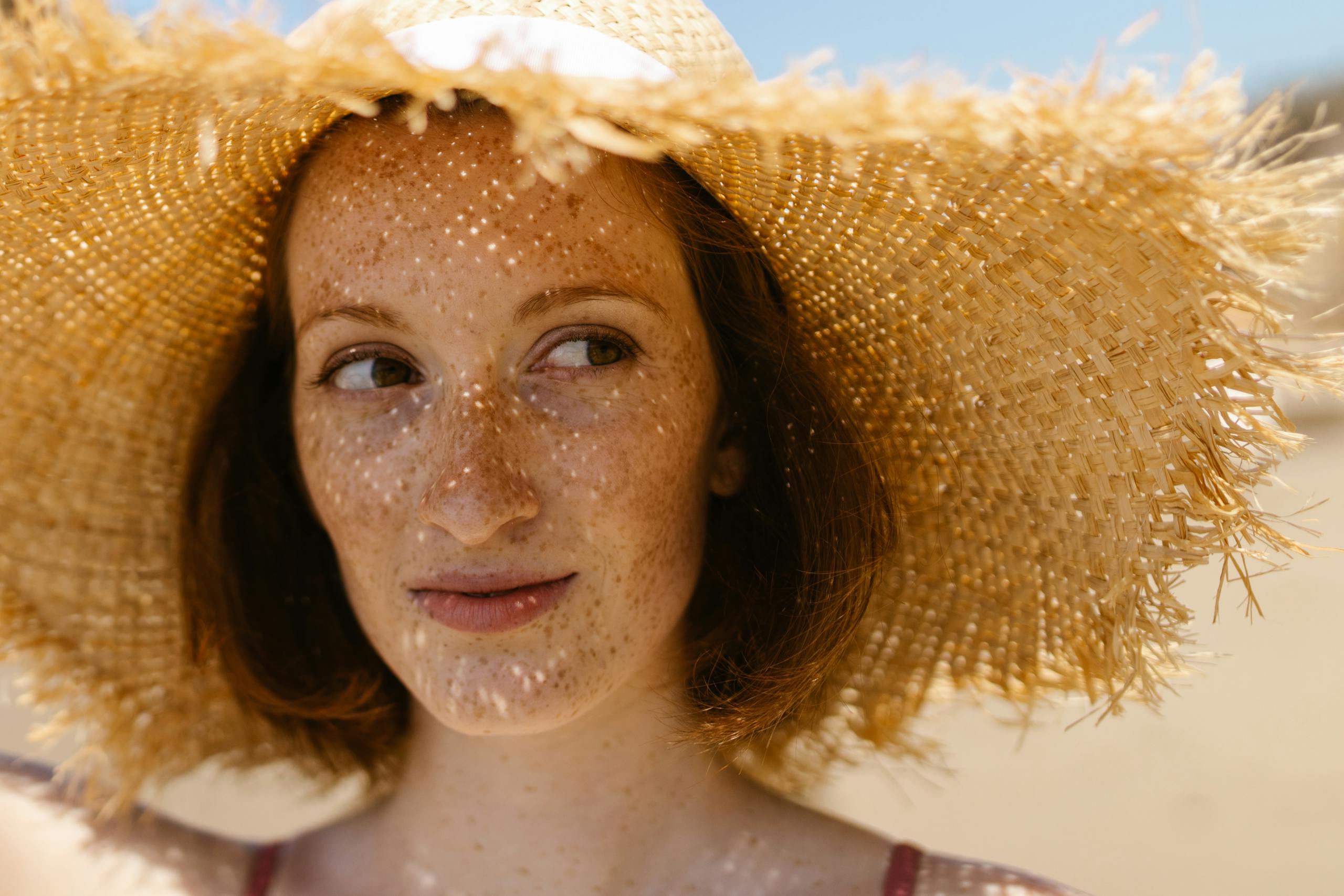 Close-up of a smiling woman with freckles wearing a straw hat on a sunny beach.