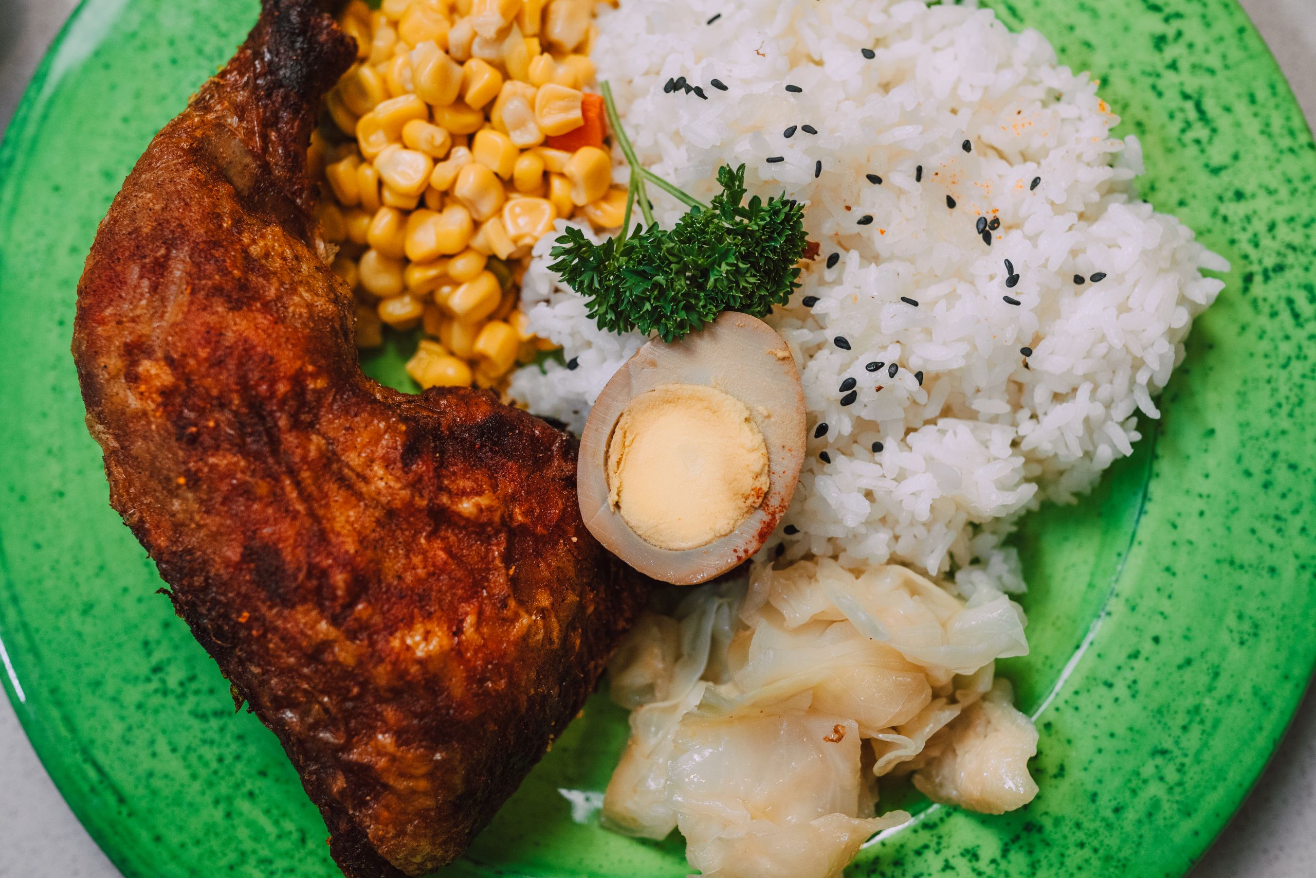 Close-up of a tasty chicken meal with rice, corn, egg, and cabbage on a green plate, styled appetizingly.