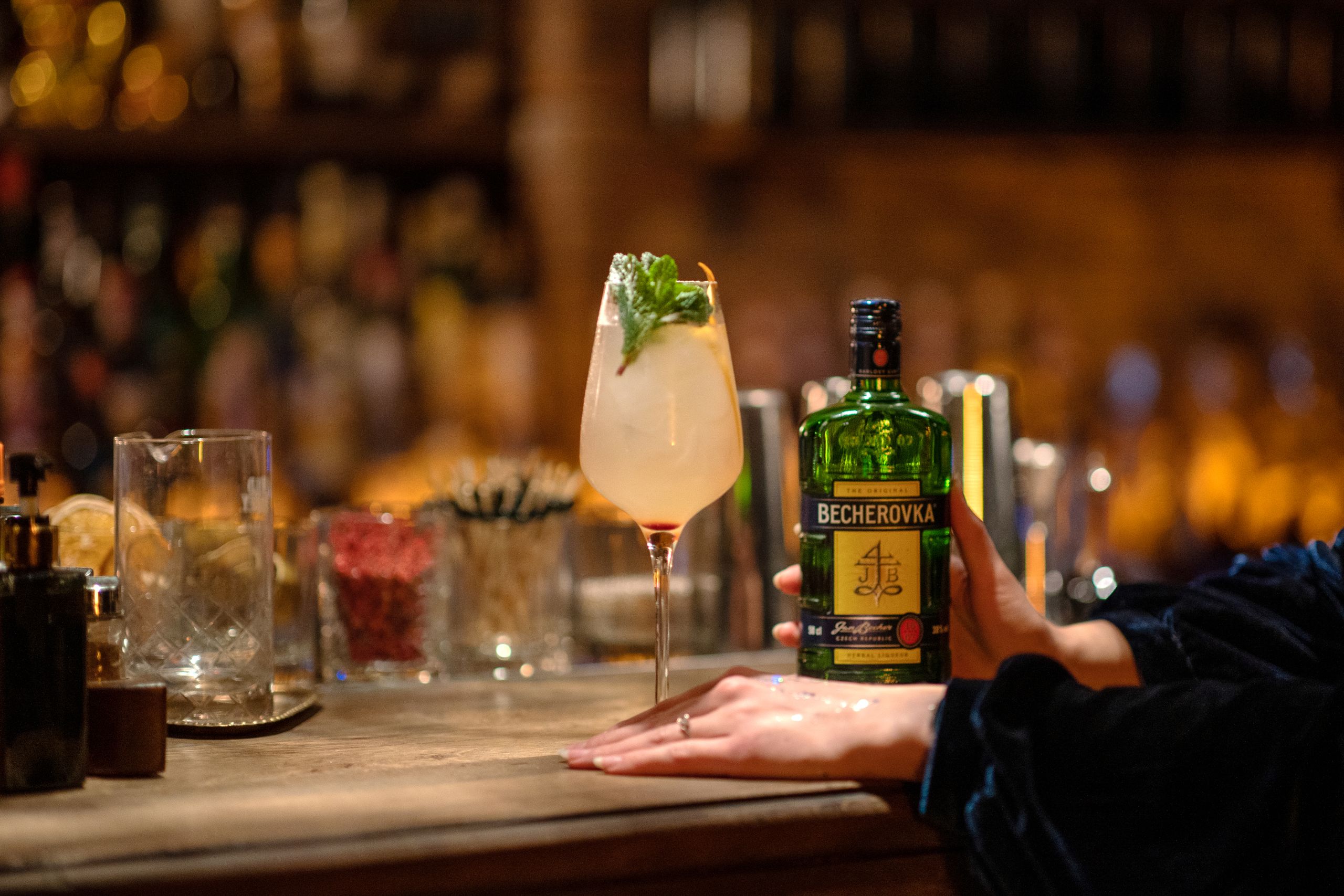 Close-up of Becherovka liquor bottle and cocktail garnished with mint at a stylish bar setting.