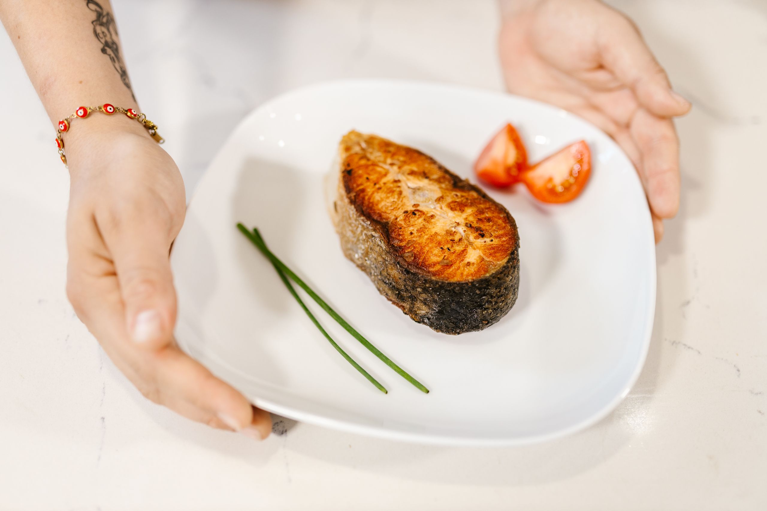 Close-up of grilled salmon served with tomatoes and chives on a plate, held by tattooed hands.