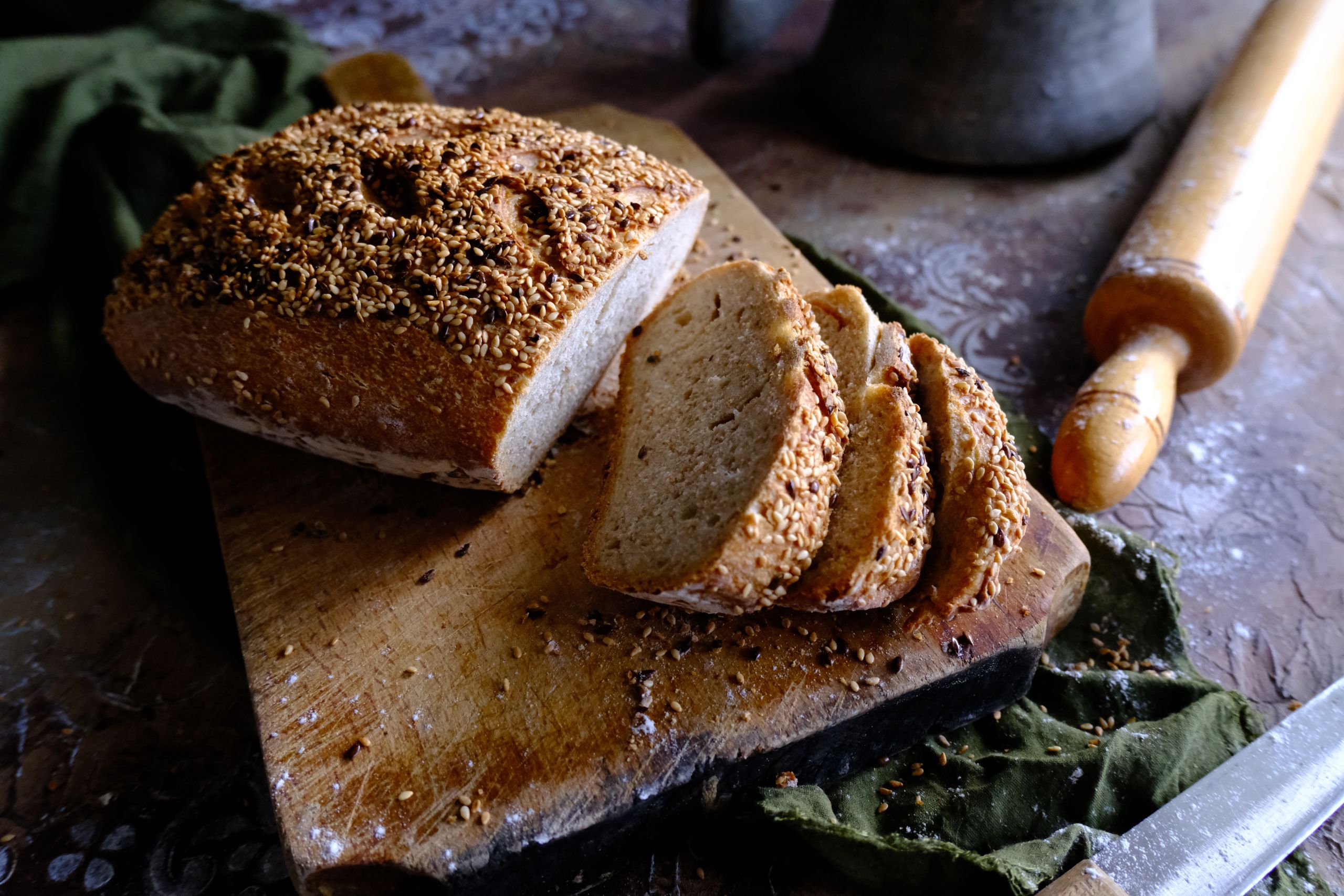 Close-up of rustic seed bread slices on cutting board with rolling pin and knife.