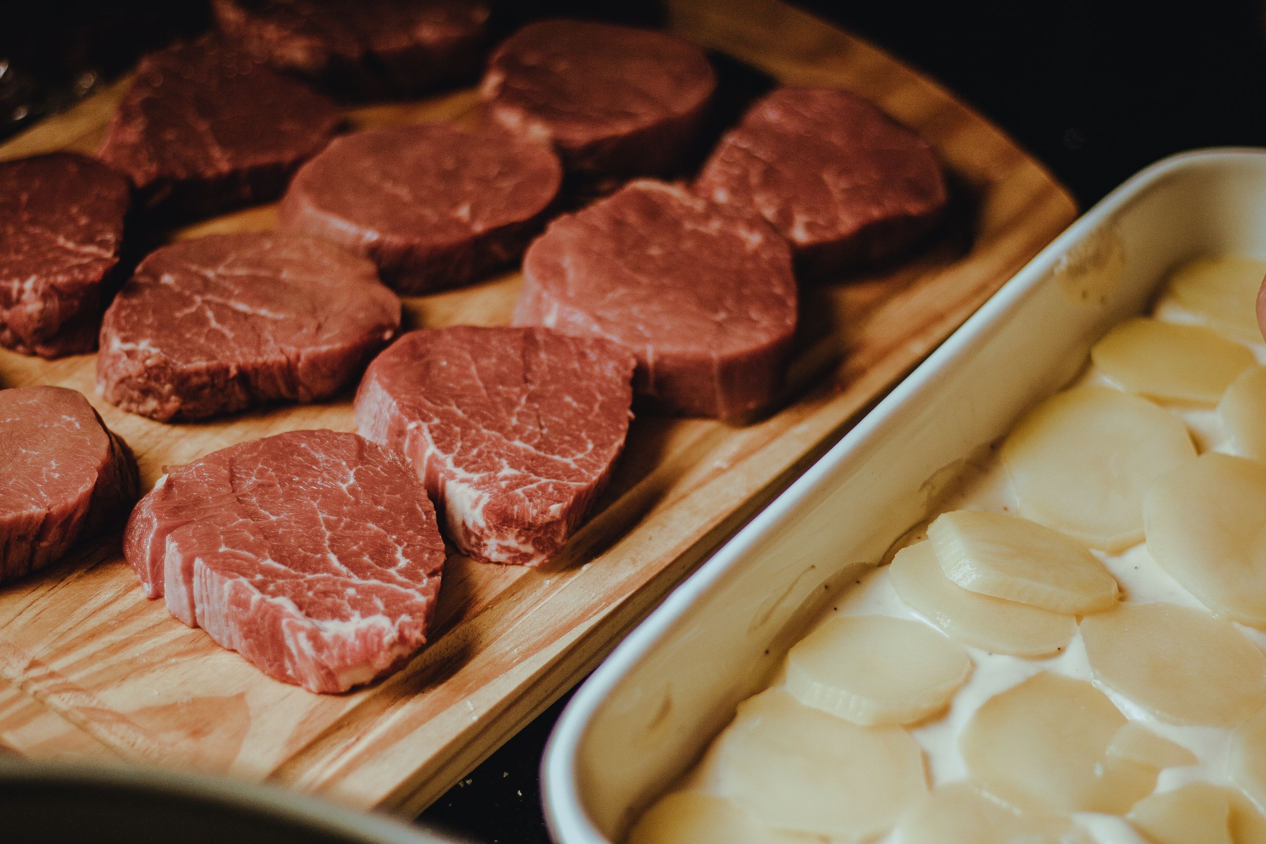 Close-up of uncooked beef steaks and potato slices on a rustic wooden tray, perfect for a gourmet meal.